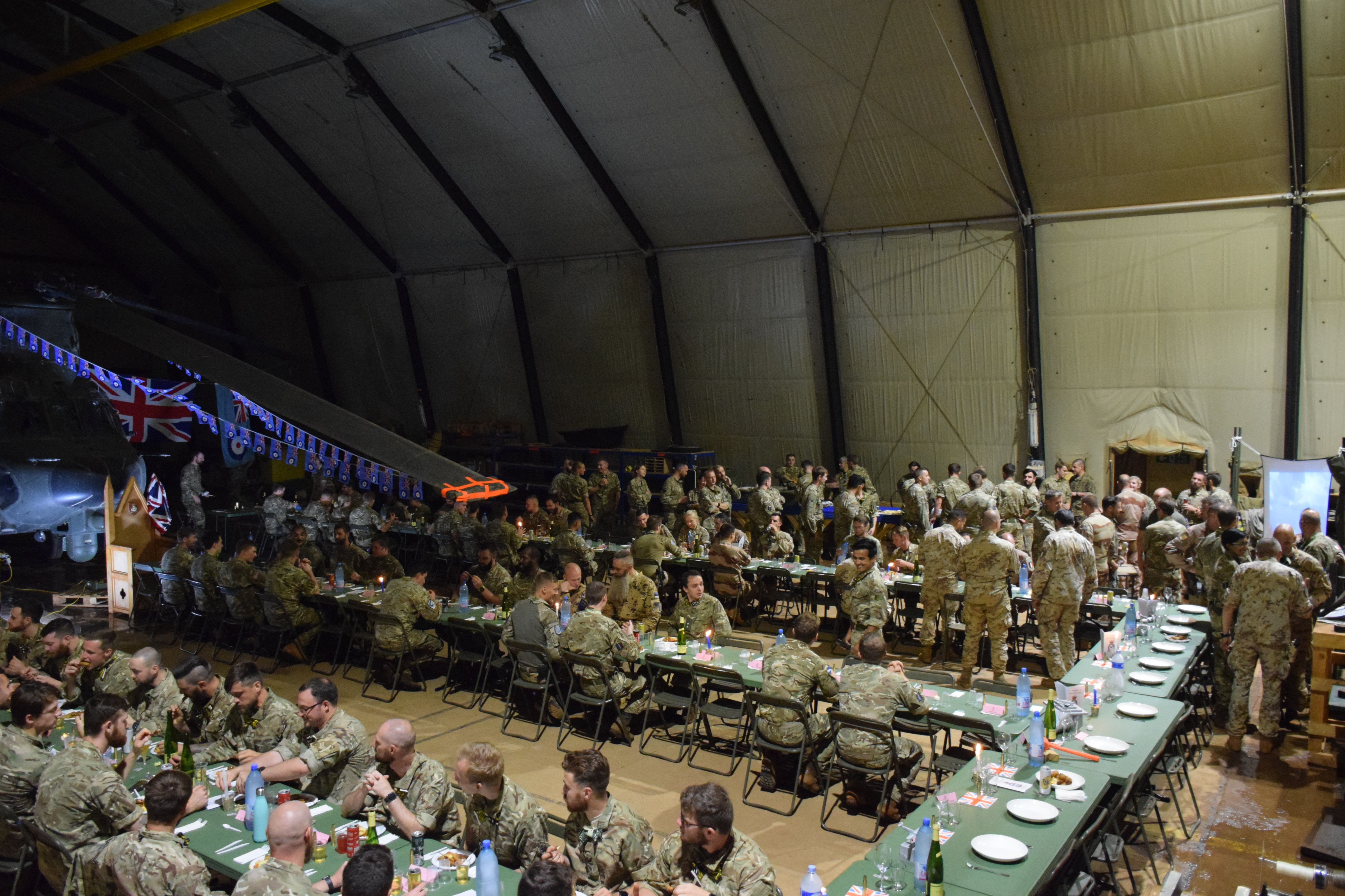Personnel eating at row of tables inside a hangar, with a Chinook as a backdrop.